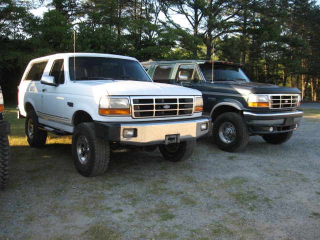 White and dark green Ford Broncos parked side by side on gravel, lifted on all-terrain tires