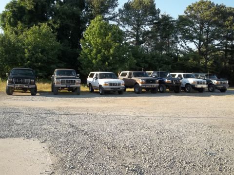 1995 Ford Bronco - A row of Ford Broncos and F-Series trucks are parked in a gravel lot with trees in the background.