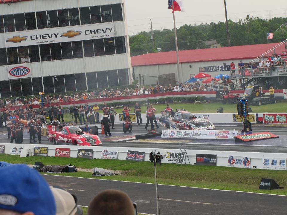 two drag cars lined up at starting line, crowd watching from stands, Chevy building in background