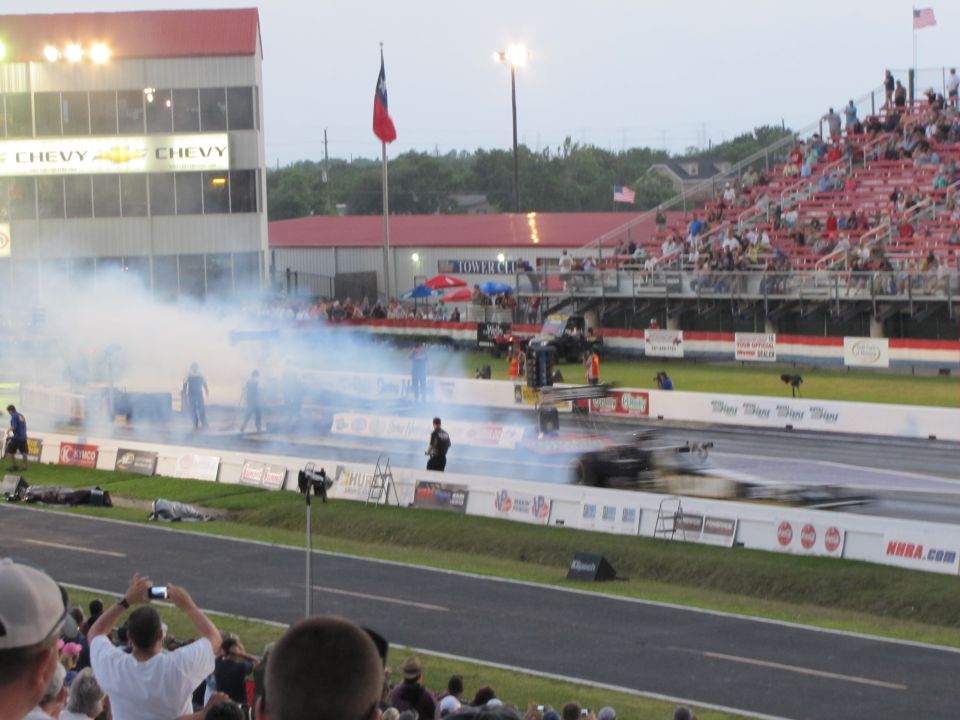 Drag strip with smoke, spectators in bleachers, and a blurred race car in motion