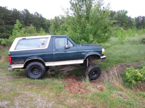 1992 Ford Bronco - A lifted 1992 Ford Bronco with a dark green body and white top is shown from the side in a grassy, wooded area.