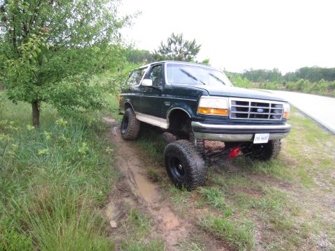 1992 Ford Bronco - A lifted green 1992 Ford Bronco is parked on a muddy dirt path next to a tree.
