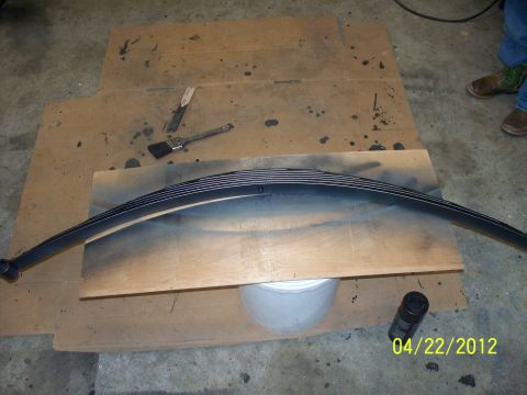 1990 Ford Bronco - A leaf spring is being painted black on a wooden board, with tools and a spray can nearby.