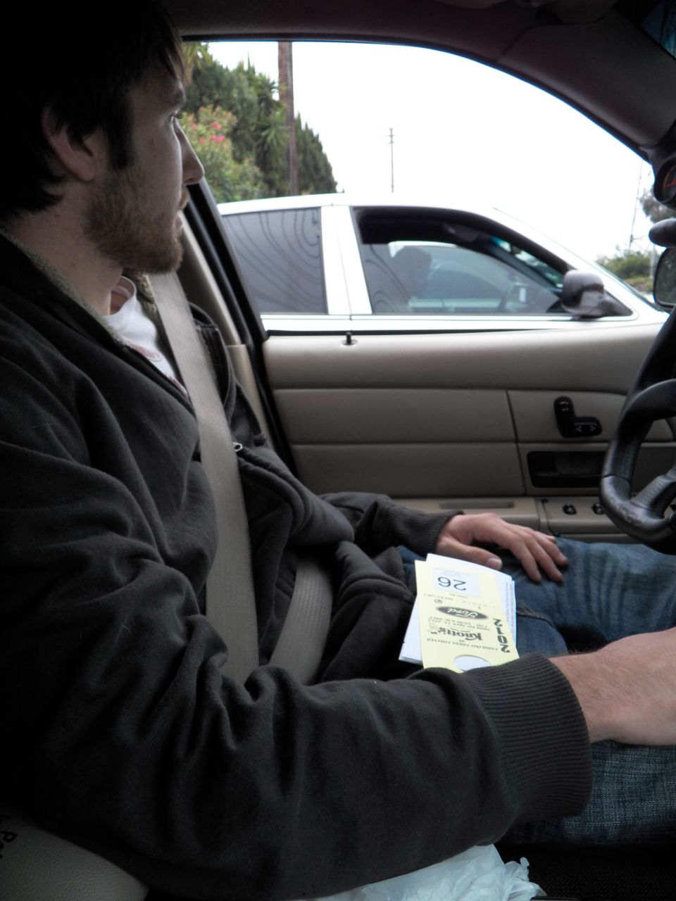 Man in dark hoodie seated in passenger seat of sedan holding event flyers numbered 26