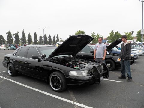 2003 Ford Crown Victoria - Black Crown Victoria on black wheels with raised hood; bystanders chat under gray skies.