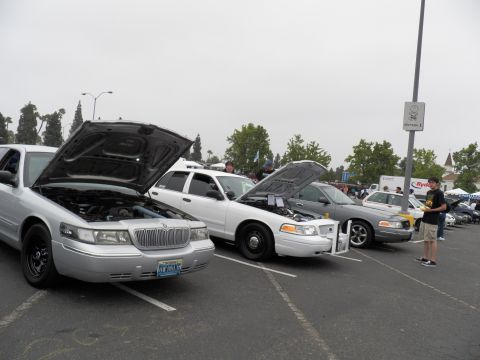 2003 Ford Crown Victoria - Silver Mercury sedan and white Ford parked with hoods raised; an attendee reads a flyer nearby.
