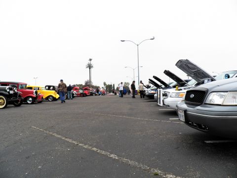 2003 Ford Crown Victoria - Gray Ford sedan front at right; classic hot rods and a crowd fill the lot at left