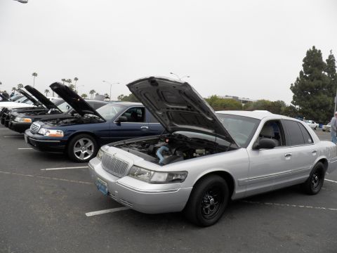 2003 Ford Crown Victoria - Silver Mercury sedan with raised hood at right; blue and dark sedans line the row at left