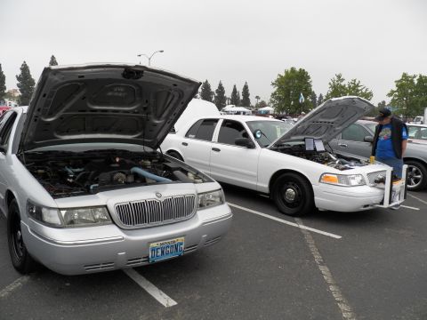 2003 Ford Crown Victoria - Two full-size sedans with raised hoods; silver Mercury at left, white Ford with push bar at right