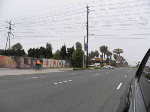 2003 Ford Crown Victoria - Bicyclist in orange jacket beside an ENJOY mural on a wide street, signage and palms in distance