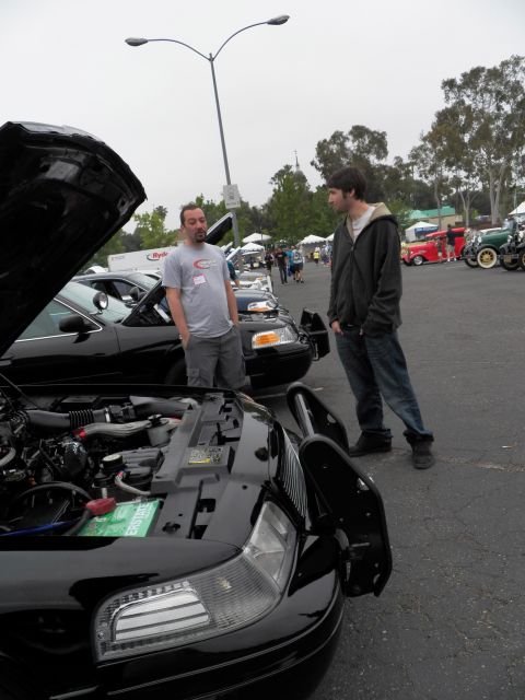 2003 Ford Crown Victoria - Bearded man in gray shirt and younger man chat over an open black engine bay; tents and a hot rod behind