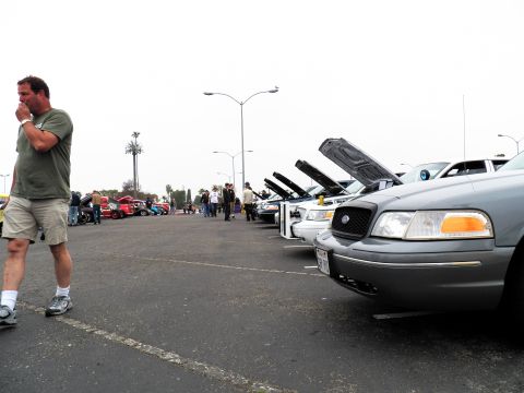 2003 Ford Crown Victoria - Spectator in green shirt and shorts strolls along a row of hood-up sedans, silver Ford nearest