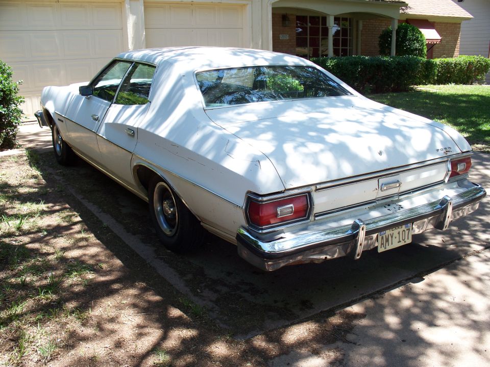 White 1977 Ford sedan, rear 3/4 view, parked in driveway. Chrome bumper, rectangular taillights