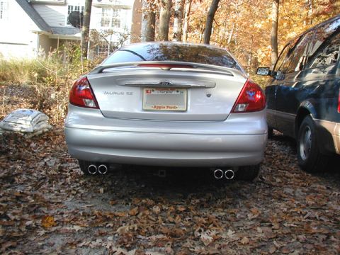2000 Ford Taurus - Straight-on rear view of a silver Ford Taurus SE sedan parked on a leaf-strewn driveway, showing the decklid spoiler, TAURUS SE badge and a…