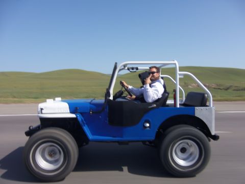 1968 Ford Bronco - Man drives a blue and white jeep on a road with rolling hills under a clear blue sky.