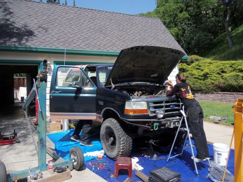 1968 Ford Bronco - A person works on the engine of a Ford F-Series truck in a driveway, with a garage and house in the background.