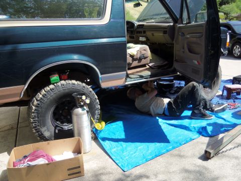 1968 Ford Bronco - Mechanic works on the underside of a Ford Bronco, with tools and equipment laid out on a blue tarp.