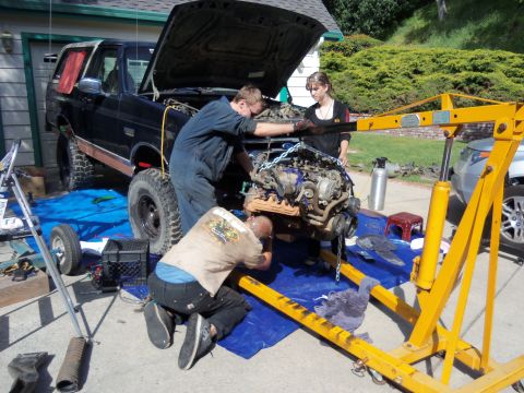 1968 Ford Bronco - Mechanics work on removing the engine from a black SUV using an engine hoist.