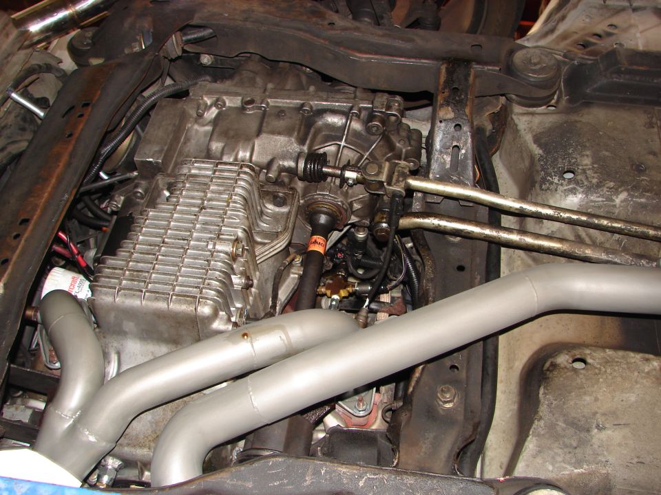 Underside view of a car's transmission, oil pan, and exhaust pipes. Some wiring and hoses are visible