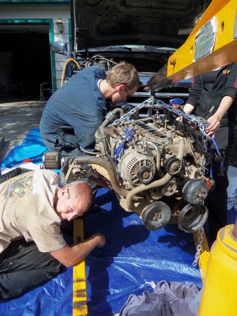 1968 Ford Bronco - Mechanics work together to remove an engine from a vehicle using an engine hoist, with a blue tarp spread underneath.