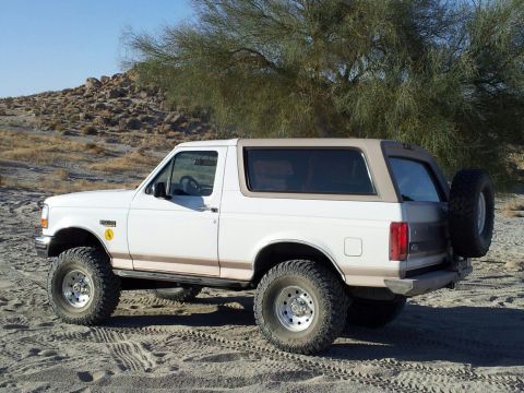 1996 Ford Bronco - White 1996 Ford Bronco with tan top and spare tire on the back, parked in a sandy area.
