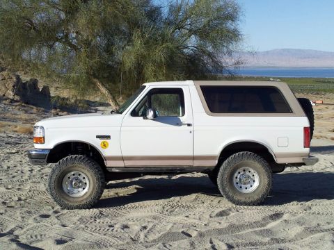 1996 Ford Bronco - This 1996 Ford Bronco is shown in profile, parked on a sandy surface with a tree and body of water visible in the distance.
