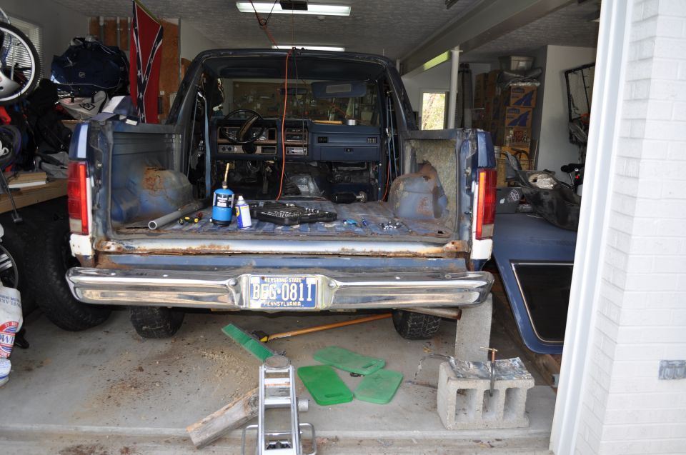 Rear view of a blue Ford Bronco in a garage, interior disassembled. Tools and a propane torch are on the bed