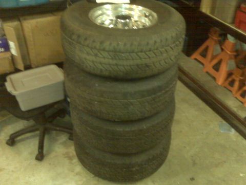 1995 Ford Bronco - A stack of four tires with a chrome rim on the top tire is pictured. The image also shows a chair and boxes in the background.
