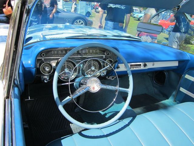 Interior view of a classic car with a light blue dashboard and steering wheel, showing gauges and seats. Other cars are visible outside