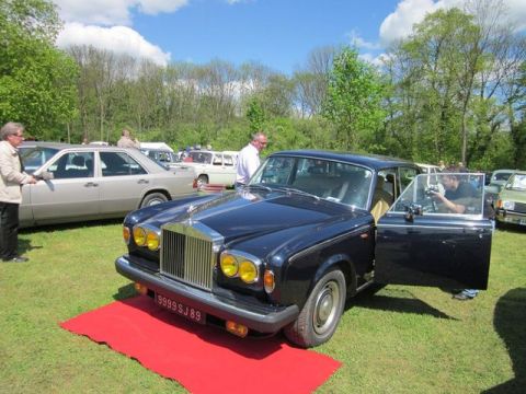 1991 Ford LTD Crown Victoria - A dark blue Rolls-Royce Silver Shadow is displayed on a red carpet at an outdoor car show, with other vehicles visible in the background.