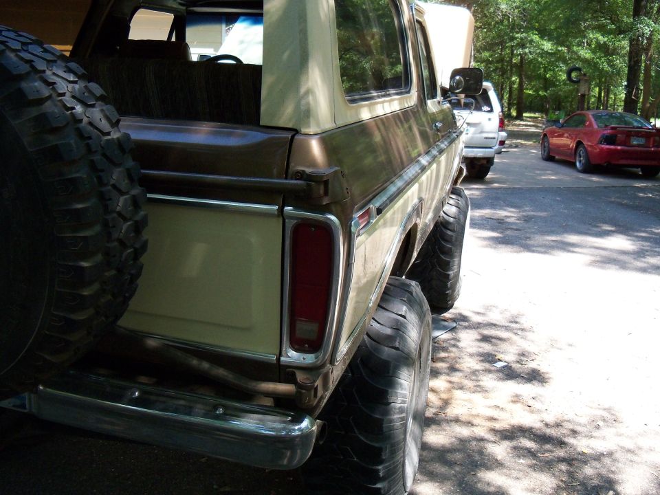 Rear view of a lifted Ford Bronco with a spare tire on the back. A white truck and a red car are parked behind it