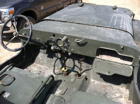 1986 Ford Bronco - The interior of a weathered olive drab military jeep is shown, with a dark pickup truck in the background.