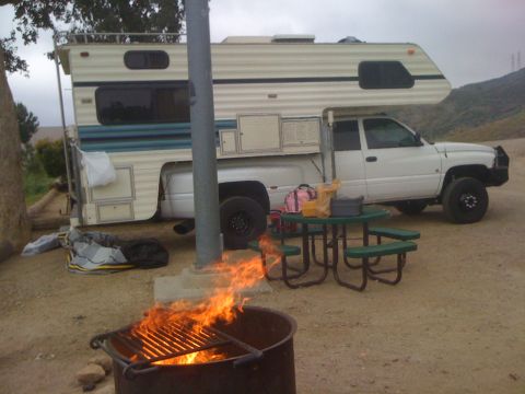 1997 Dodge 3500 - A white pickup truck with a camper shell is parked at a campsite with a fire pit in the foreground.