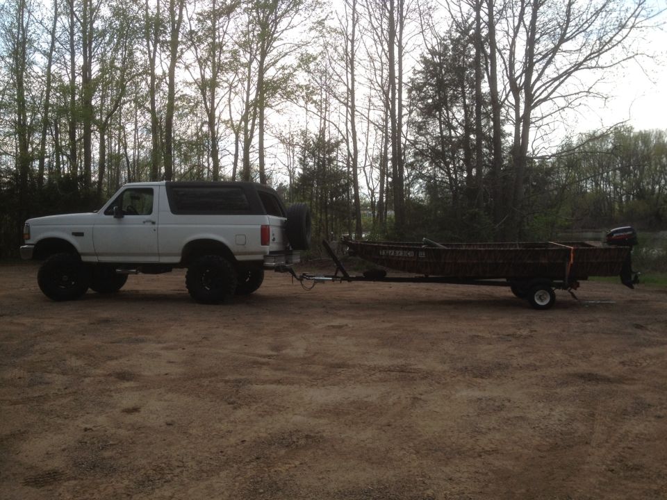 White 1992 Ford Bronco with lifted suspension and large tires, towing a camouflage boat trailer