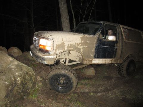 1992 Ford Bronco - Muddy 1992 Ford Bronco tackling rough terrain on a dark, wooded trail.
