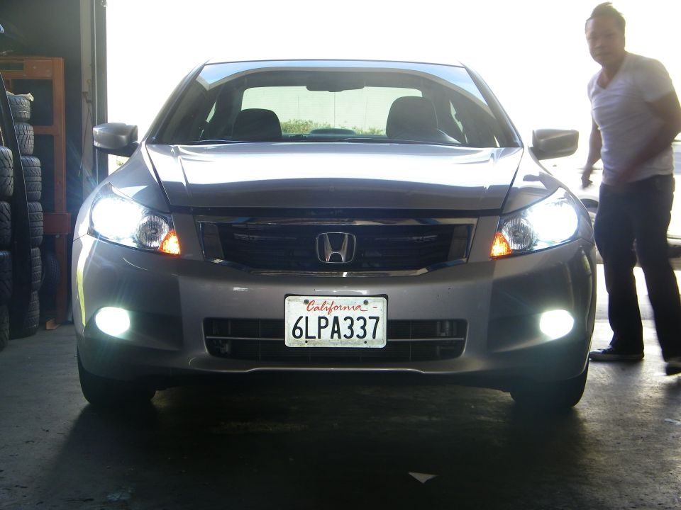 Front view of a silver Honda Accord with its headlights and fog lights on. A person stands to the right