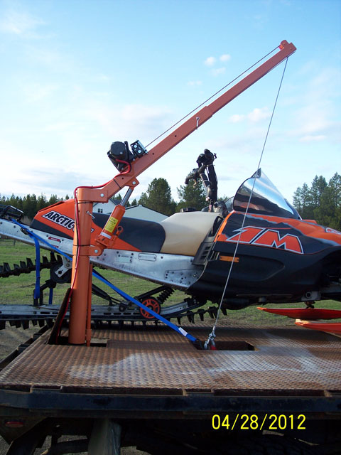 1973 Ford F-250 - An orange crane is positioned over a snowmobile that is strapped to a flatbed trailer.