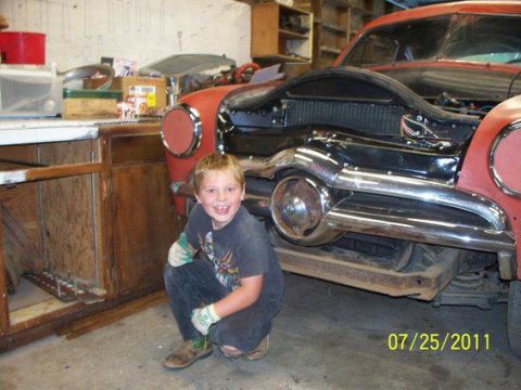 1990 Ford Bronco - A young boy poses in front of a vintage car undergoing restoration in a garage.