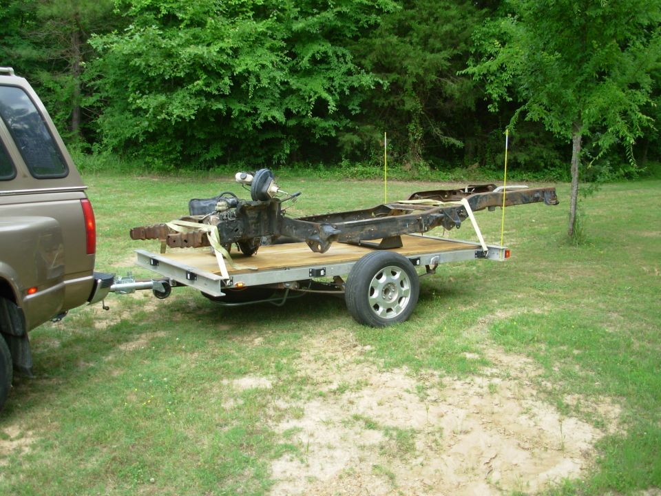 Utility trailer hauling a bare rusty truck frame behind a gold SUV on grass