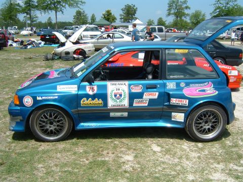 1997 Ford Taurus - A blue hatchback covered in various racing decals is parked on a grassy field with other vehicles in the background.