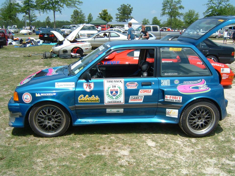 Blue hatchback with racing stickers and aftermarket wheels parked on grass