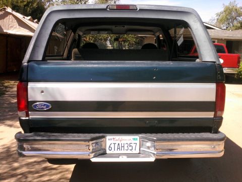 1994 Ford Bronco - Rear view of a 1994 Ford Bronco, showing its tailgate and bumper. A red truck is partially visible.