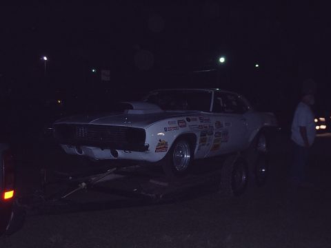 1996 Ford Bronco - A white drag car covered in racing decals is parked on a trailer at night.