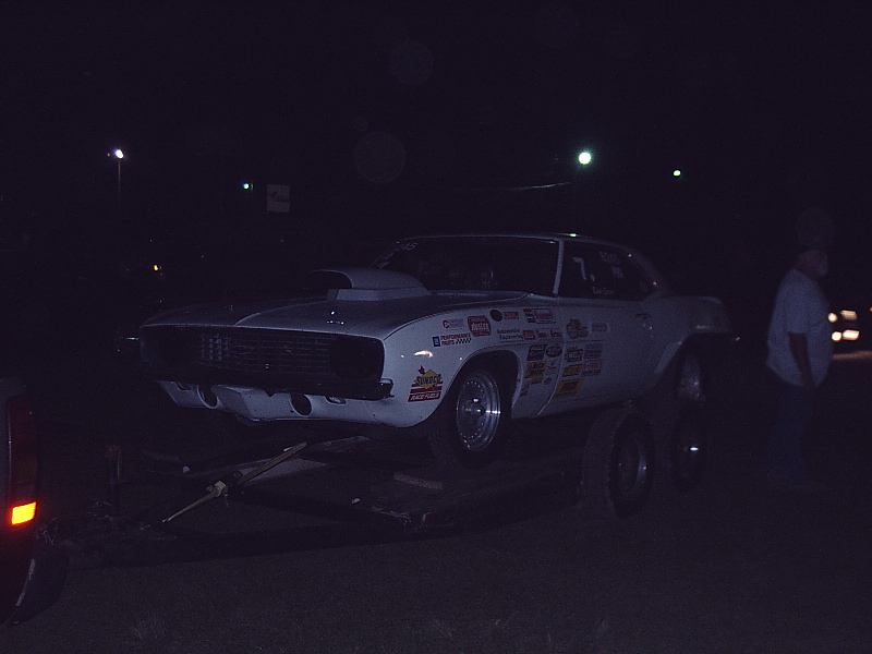 White Camaro drag car with sponsor decals loaded on a trailer at night