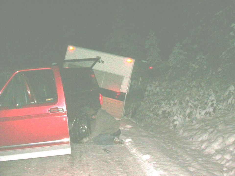 Red pickup truck with trailer stuck in snow, person working on wheel