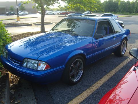 2007 Ford Mustang - A blue Ford Mustang from the late 1980s is parked in a lot with other vehicles visible in the background.