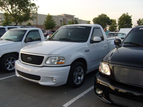 2007 Ford Mustang - A white Ford F-150 Lightning pickup truck is parked in a lot with other vehicles, with a custom exhaust banner on its windshield.