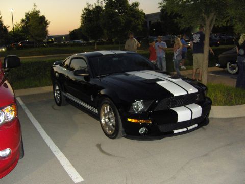 2007 Ford Mustang - A black 2007 Ford Mustang with white racing stripes is parked in a lot with other vehicles and people around.