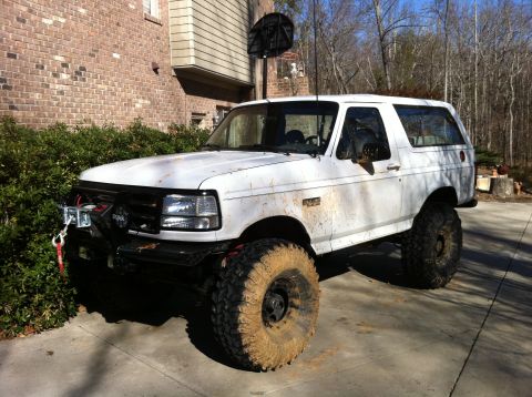 1996 Ford Bronco - Three-quarter front view of a lifted white full-size Ford Bronco caked in dried mud, wearing tall mud-terrain tires on black steel wheels a…