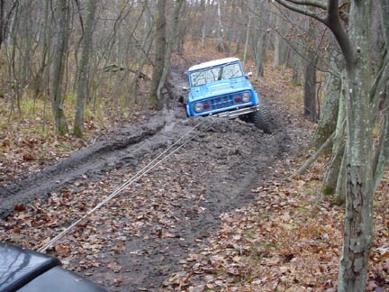 1989 Ford Bronco - A blue early Ford Bronco with a white roof sits nose-down at the top of a rutted, muddy trail climb in leafless woods, with a winch cable r…